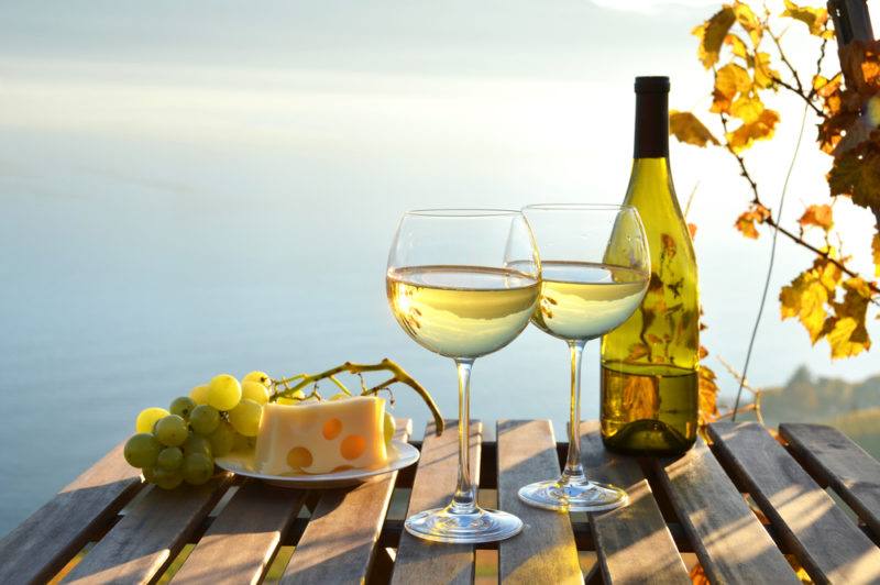 A bottle of white wine with two glasses of white wine outside next to Swiss cheese and grapes