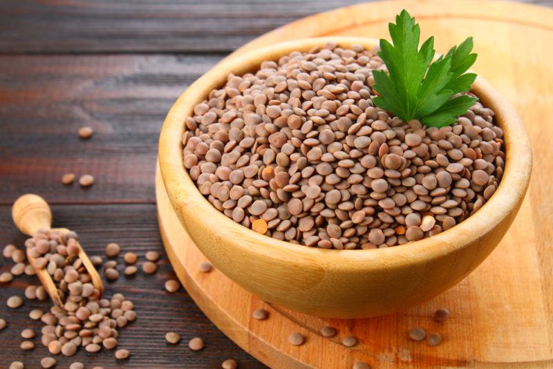 A light brown bowl on a wooden board that contains raw lentils, next to a wooden scoop with more lentils