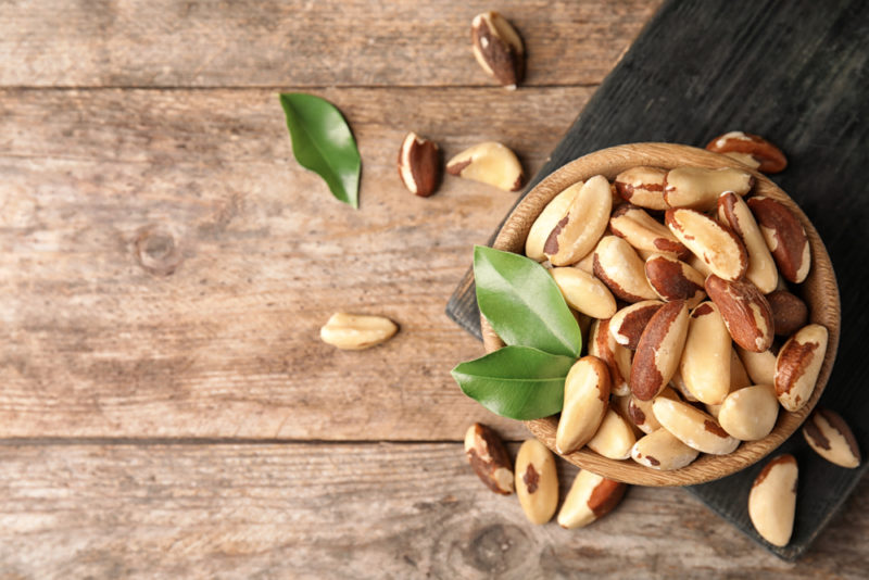 A bowl of Brazil nuts on dark board on a wooden table with a few Brazil nuts scattered around