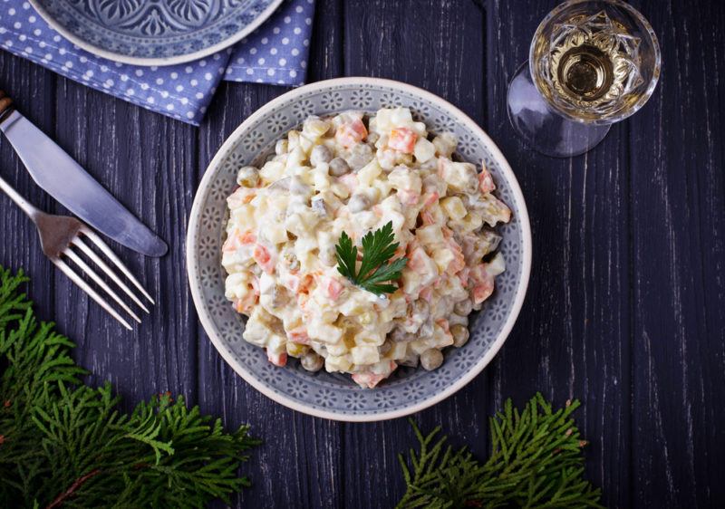 A bowl of Olivier salad on a wooden table, next to a knife, fork, and some greenery