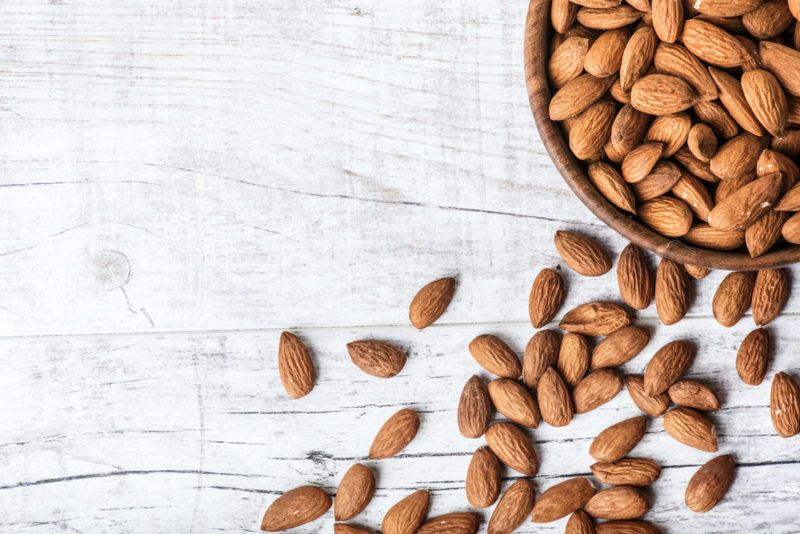 A brown bowl filled with almonds, with more almonds on a wooden table