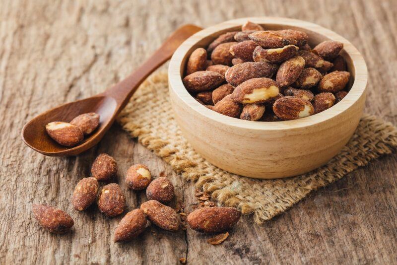 A bowl of salted roasted almonds on a cloth, with more almonds on the table