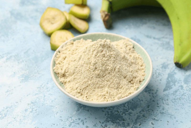 A light colored table with a white bowl of banana flour, sliced bananas, and a green banana.