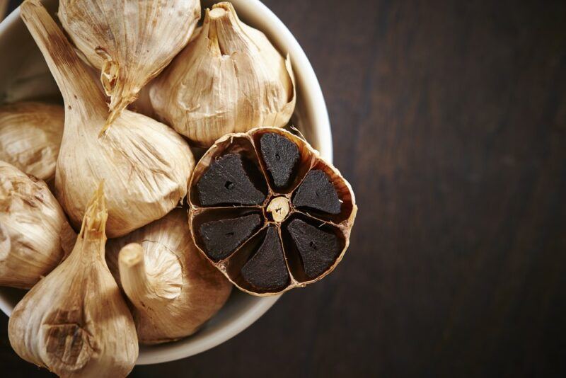 A bowl filled with black garlic. One of the garlic bulbs has been cut open so we can see the black garlic cloves