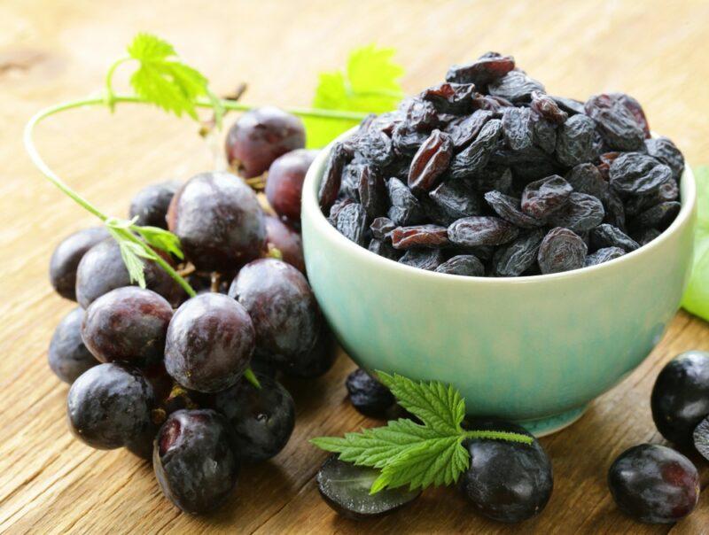 A small white bowl of raisins next to some fresh black grapes