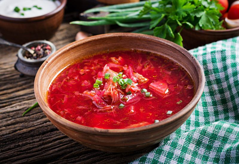 A brown bowl of borscht that is colored red from the beets, next to a green and white cloth and in front of some vegetables