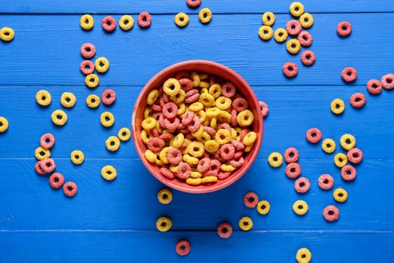 A dark blue wooden table with a bowl of pink and yellow breakfast cereal circles, plus more of the cereal scattered on the table