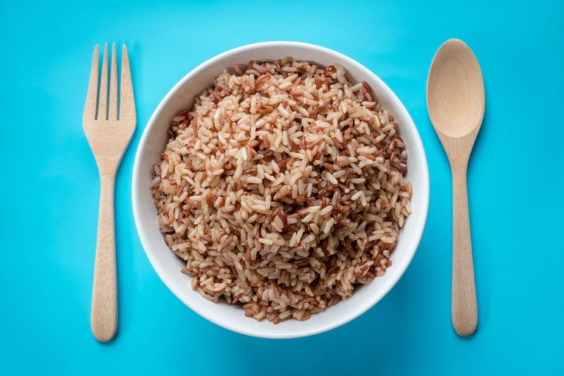 A white bowl of brown rice, with a wooden knife and fork