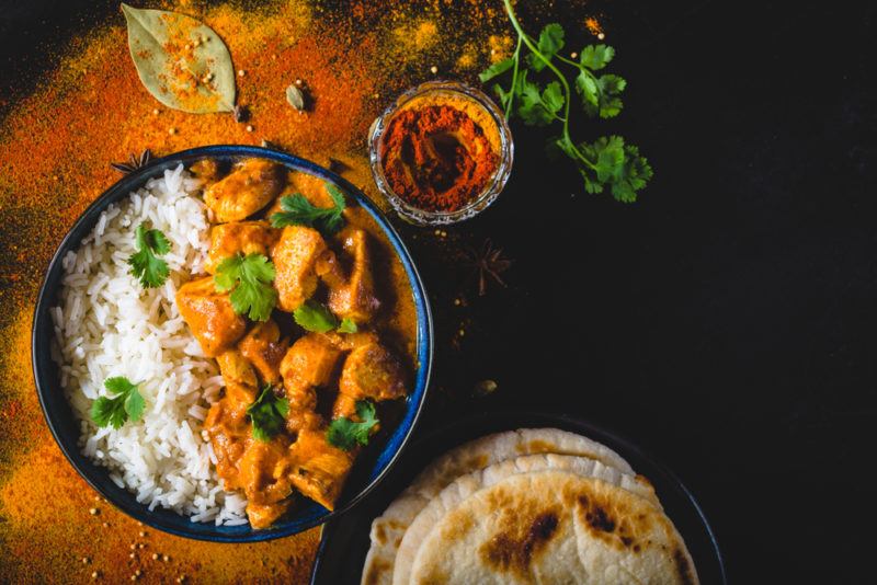 A black table with a bowl of butter chicken and rice, next to naan bread and spices