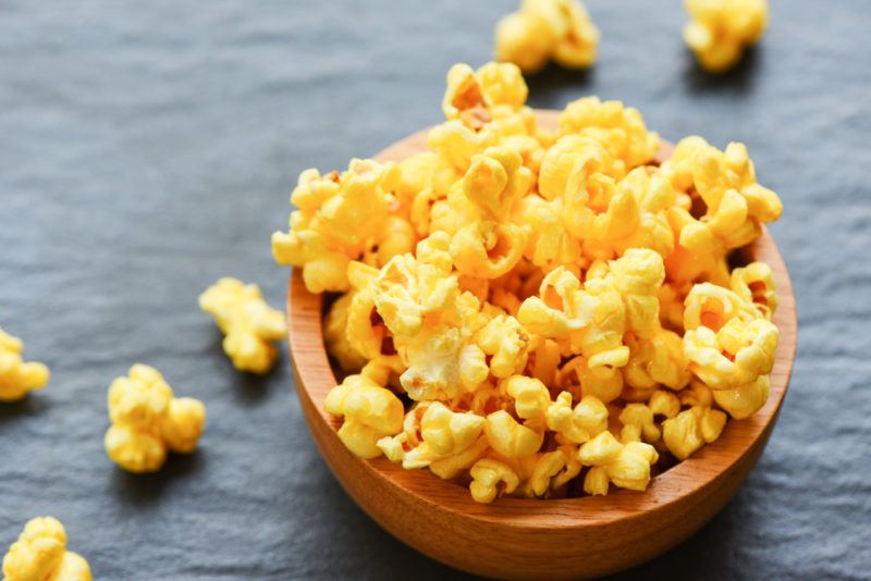 A wooden bowl of buttered popcorn with a few pieces scattered on the table