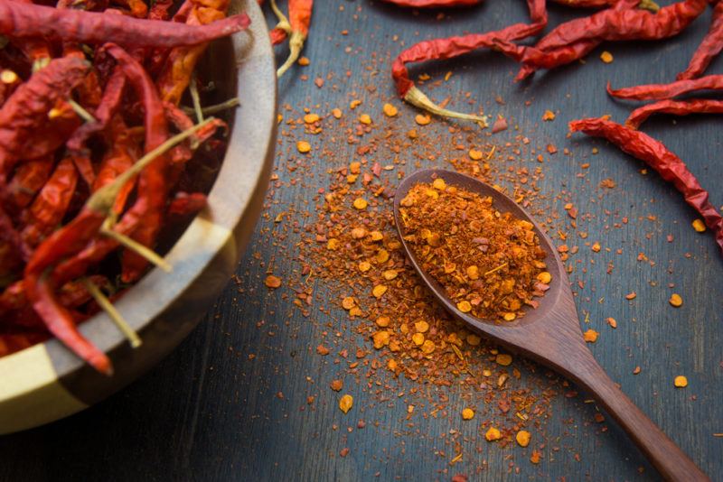 A wooden bowl with cayenne peppers, with a spoonful of the ground peppers and a few on the table