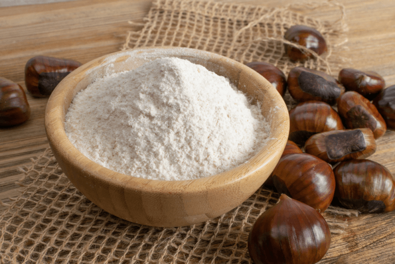 A wooden table with a bowl of chestnut flour, plus chestnuts scattered on it.