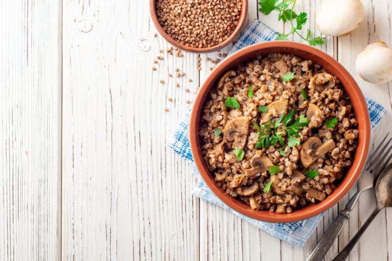 A red brown bowl containing buckwheat porridge with mushrooms, next to a bowl of buckwheat groats