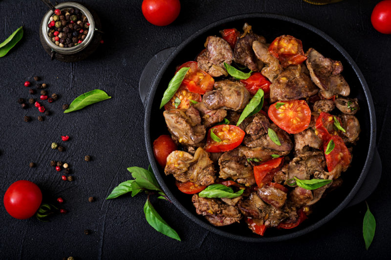 A black bowl of cooked chicken liver with tomatoes and green herbs on a black table with seasonings and more herbs