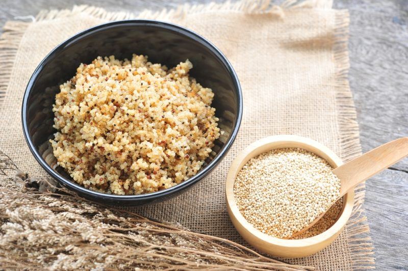 A dark gray bowl of cooked quinoa, with a smaller bowl of uncooked quinoa next to it