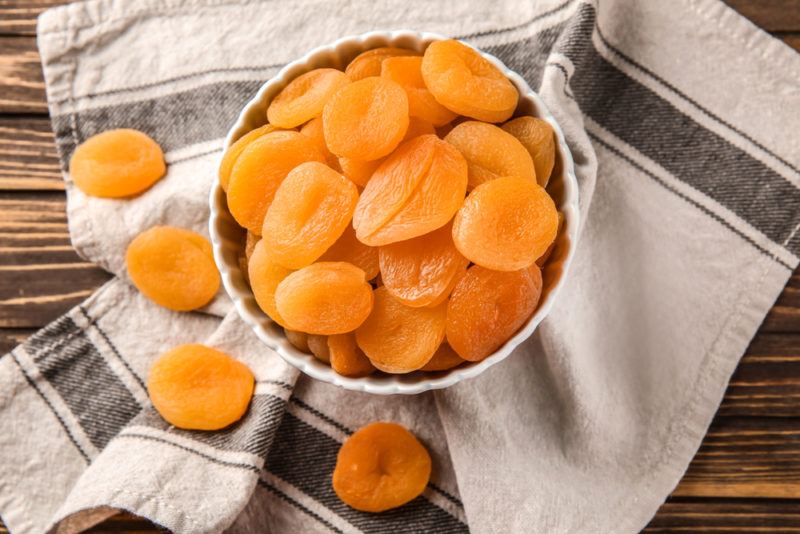 A white bowl of dried apricots on a tea towel with more apricots scattered on the towel