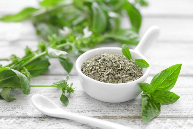 A white cup of dried basil next to fresh basil leaves