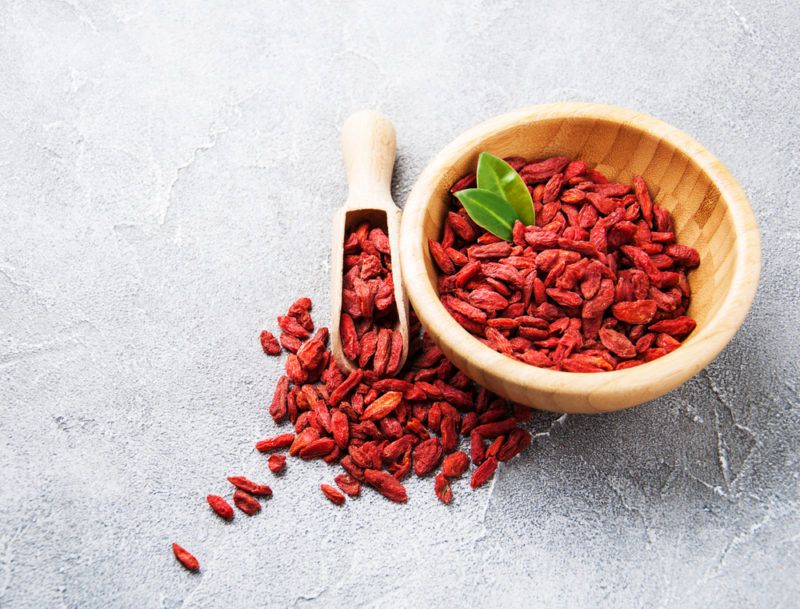 A wooden bowl partly filled with goji berries, next to a wooden scoop and more varries scattered on the table