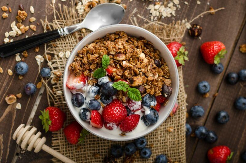 A white bowl of granola, yogurt, and fruit, on a wooden table