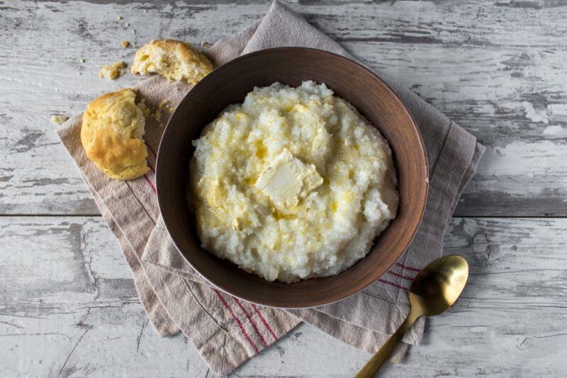 A brown bowl of grits with butter on a wooden table, next to a spoon