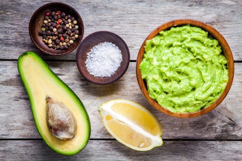 A bowl of guacamole, next to a lemon wedge, small bowls of salt and pepper, and half an avocado