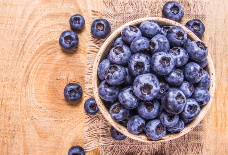 A wooden bowl of huckleberries on a wooden table and a cloth. There are also some berries on the table.