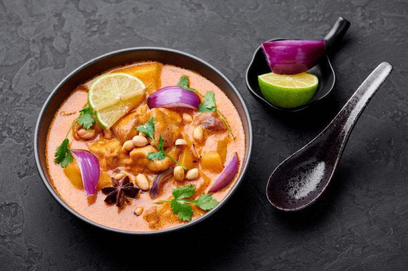 A black bowl of massaman with greens and red onion, next to a spoon and a small bowl with red onion and half a lime