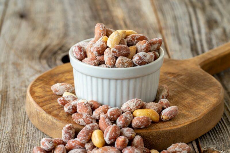 A wooden tray with a white dish of candied nuts, with more nuts spilling onto the tray