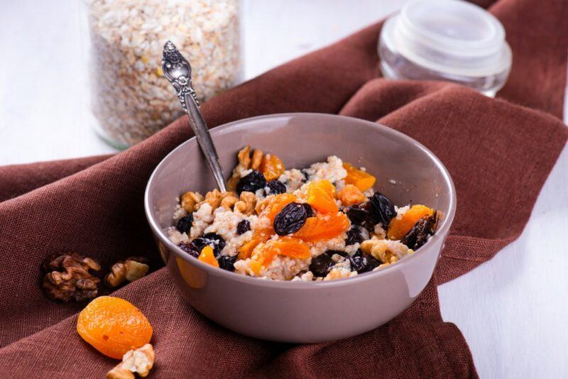 A white bowl that contains oats and dried fruit on a brown cloth, with some dried fruit scattered on it and a jar of oats