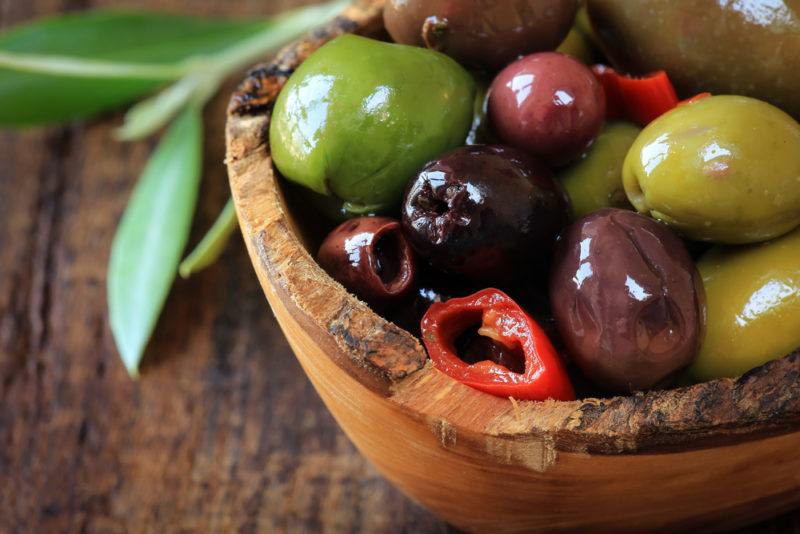 A wooden bowl containing pitted olives with brine, next to olive leaves