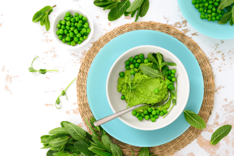 A white bowl of minted peas, some of which have been smashed, on a white table with more peas and greens nearby