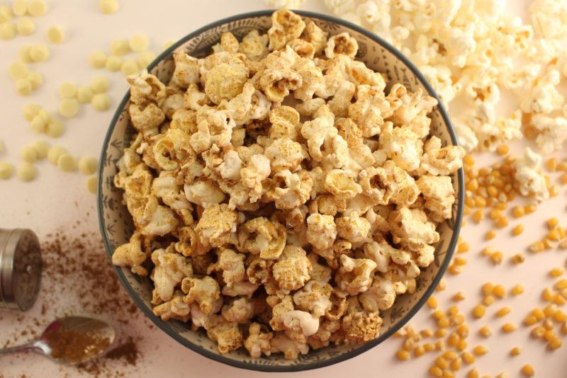 A bowl of popcorn that has been spiced, with some more popcorn and ingredients on the table