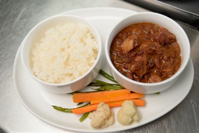 A white plate with a bowl of rice and another of chicken stew, next to a few vegetables