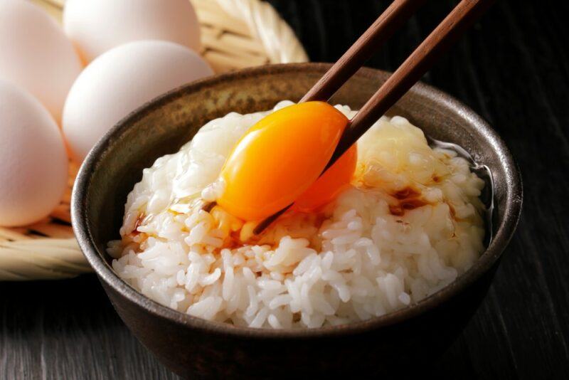 A brown bowl of rice with a raw egg that someone is grabbing using chopsticks, in front of a container of eggs