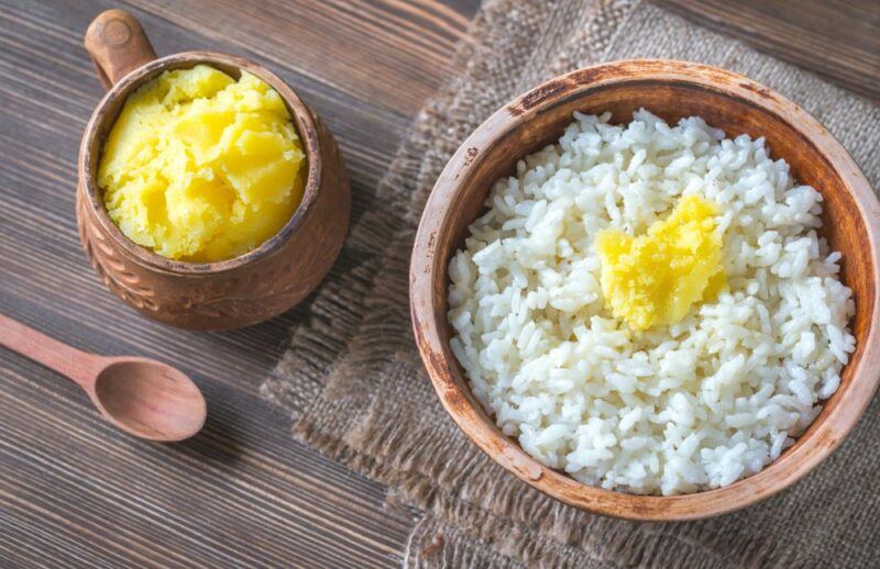 A brown bowl of rice with some ghee on top of it, near a small brown cup of ghee and a wooden spoon