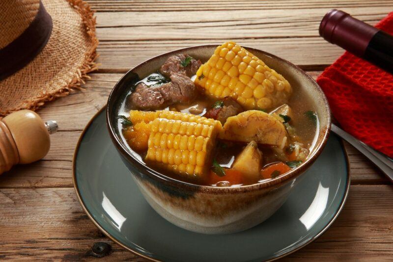 A bowl of sancoho soup with corn on the cob and plenty of meat on a plate and a table