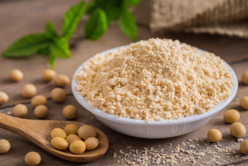 A shallow white dish of soy flour, next to a wooden spoon of soybeans and some more soybeans on a table