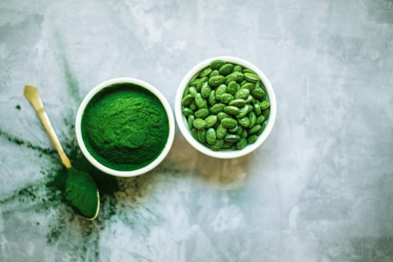 A small white bowl of spirulina and a similar bowl containing spirulina capsules. There is also a spoon of the spirulina powder.