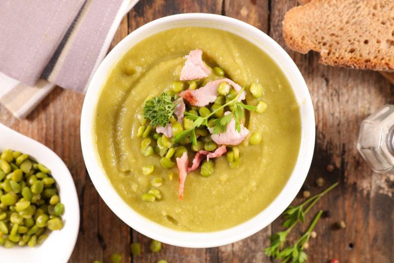 A white bowl that contains split pea soup with garnishes, next to bread and a bowl of split peas