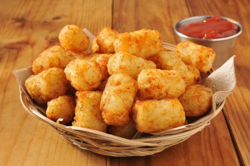 A basket with freshly cooked tater tots and a small container of tomato sauce