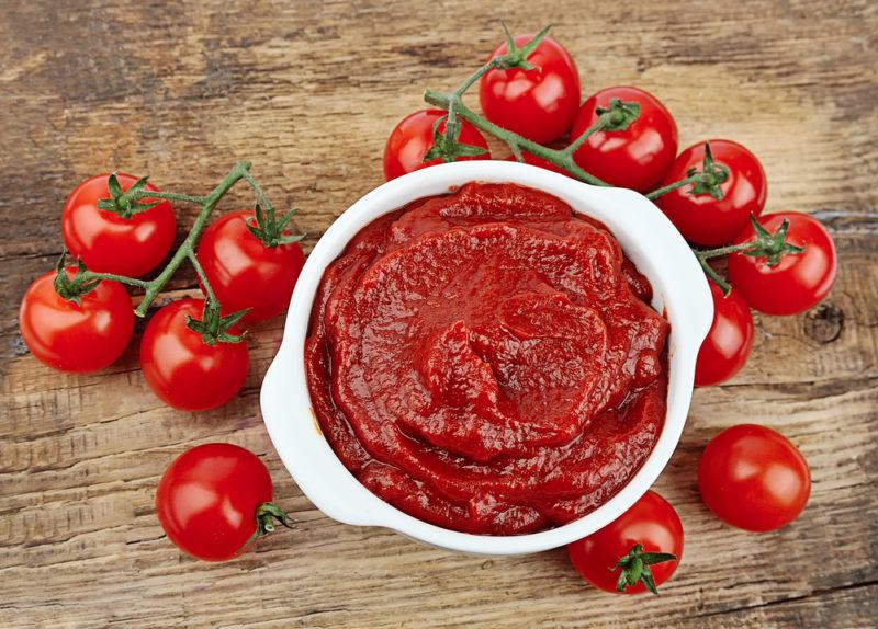 A wooden table that has a selection of fresh tomatoes, along with a white bowl of tomato paste