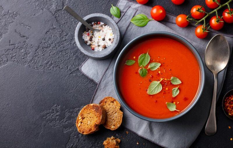 A bowl of tomato soup on a black table, near some tomatoes, seasoning, and bread