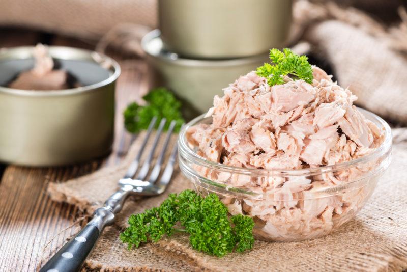 A glass bowl of tuna on a cutting board next to a fork and a can of tuna