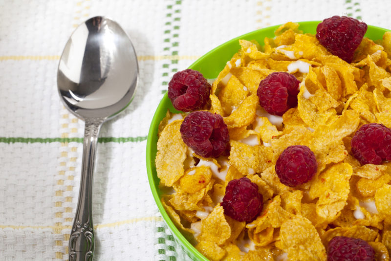 A green bowl with vitamin D fortified cornflakes, milk, and berries, next to a spoon on a tablecloth