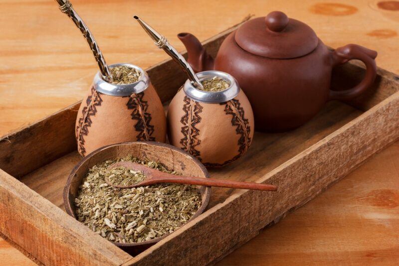 A wooden tray with two yerba mate goards, a brown bowl of the tea leaves, and a brown tea pot