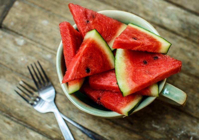 A mug of watermelon pieces on a wooden table next to two forks