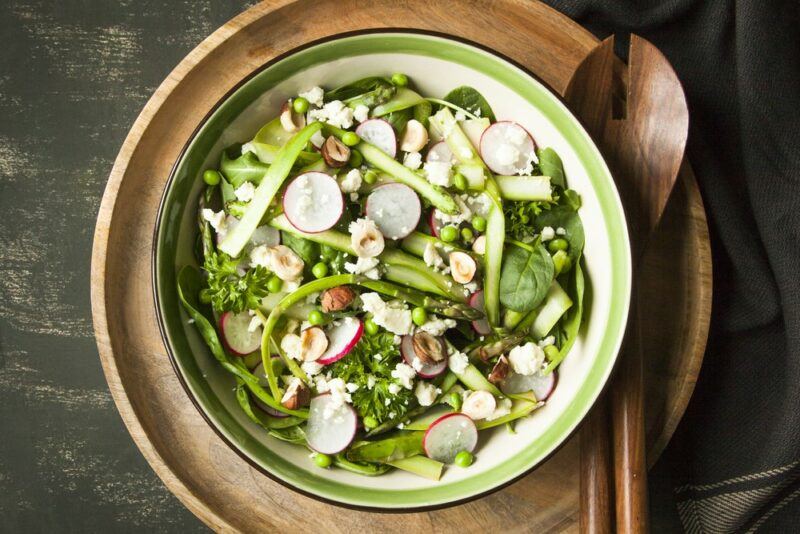 A wooden plate with a green and white dish containing a salad that includes radish slices, asparagus strips, peas, and nuts