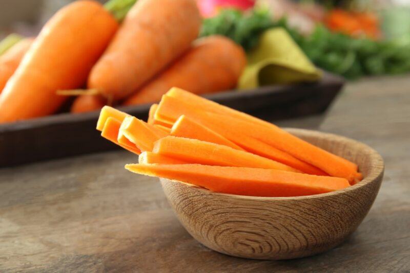 A small wooden bowl with carrot sticks on a table, with a large black dish of carrots in the background