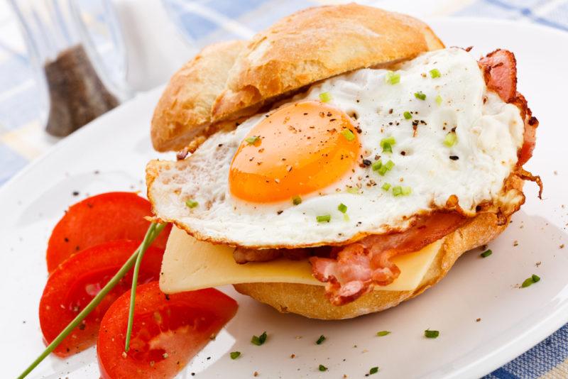 A breakfast roll on a white plate, with eggs, cheese, bacon and tomatoes