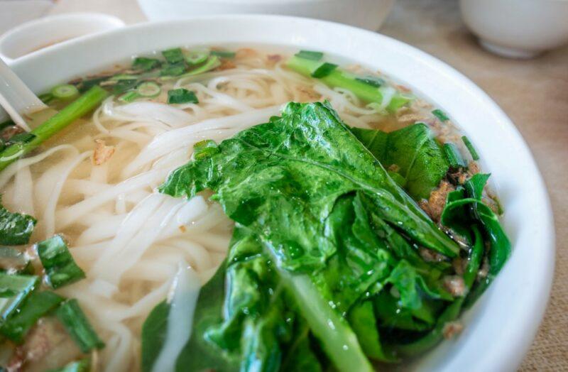 A large white bowl containing rice noodles, broth, and leafy greens